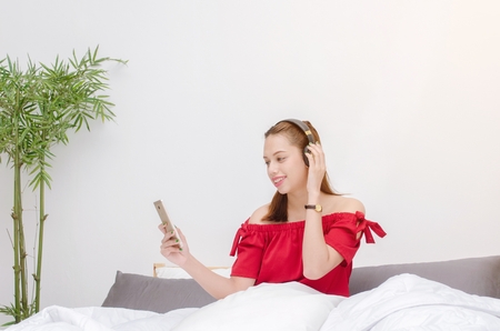young asian smiling woman in red dress relaxing and using smartphone listening to music in headphone in bedroom at the morning, technology internet and holiday lifestyle concept, sunlight effectの写真素材