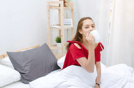 young asian woman beautiful smile in red pajamas drinking coffee on bed waking up at the morning in bedroom with sunlight effect, selective focus, soft focus, good morning, holiday, lifestyle conceptの写真素材