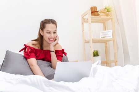 young asian beautiful smiling woman in red dress relaxing and using laptop on bed in bedroom at the morning at home, social network technology internet and holiday lifestyle concept, sunlight effectの写真素材