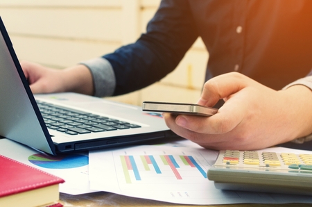 young business man using smartphone, laptop with data sheet, calculator on desk, analyzing and calculate, savings, finances, economy concept, sunlight effect, soft focus, selective focus, vintage toneの写真素材