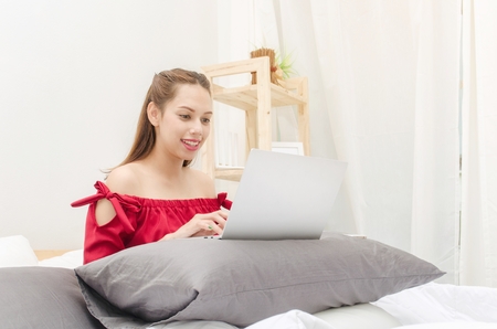 Young asian beautiful smiling woman in red dress relaxing and using laptop on bed in bedroomの写真素材