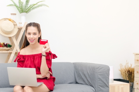 Asian young beautiful woman in red dress using laptop and sitting on sofa and holding red credit cardの写真素材