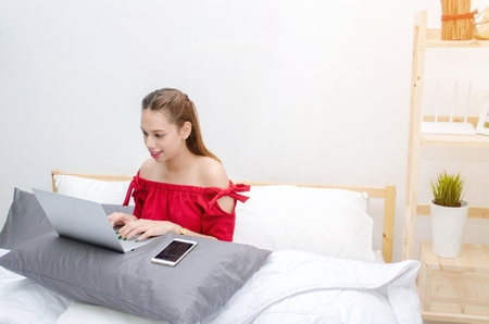 young asian beautiful woman in red dress relaxing and using laptop on in bedroom at the morning at home, social media technology internet and holiday lifestyle concept, sunlight effect, soft focusの写真素材