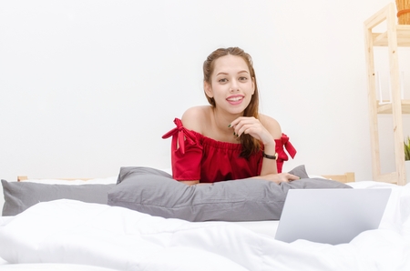 young asian beautiful woman in red dress relaxing and using laptop on in bedroom at the morning at home, social media technology internet and holiday lifestyle concept, sunlight effect, soft focusの写真素材
