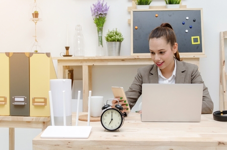 asian young business woman beautiful smile using smartphone on wooden desk with laptop computer, soft focus, sunlight effect, social network technology, business lifestyle and working woman conceptの写真素材