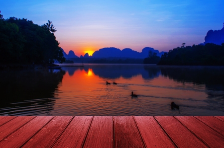 empty wooden red floor, desk or terrace copy space for display of product or object presentation with abstract blurred image of colorful sunrise mountain silhouette and duck swimming in lakeの写真素材