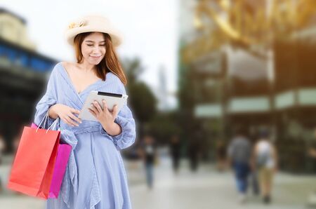 cheerful asian young woman holding shopping bags and mobile tablet while standing at the department store shopping mall, online payment, shopping online, lifestyle technology conceptの写真素材