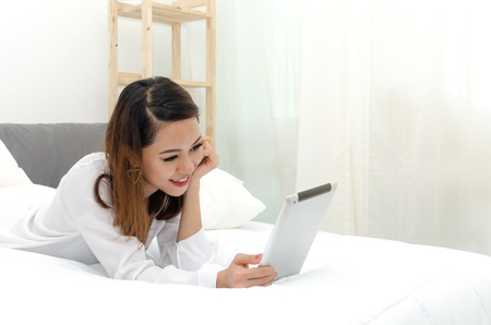 happy young asian woman in white pajamas using mobile tablet on bed in the morning at home, soft focus, lifestyle smart communication technology, online shopping and payment online conceptの写真素材