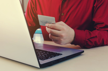 handsome man in red shirt holding credit card with laptop computer on desk,online payment, shopping online, lifestyle technology concept, selective and soft focusの写真素材