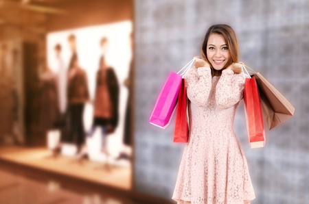 cheerful young asian woman happy smiling holding shopping bag while standing at brand name clothes shop in department store shopping mall, online payment, shopping online, lifestyle technology conceptの写真素材