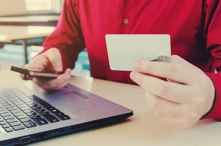 handsome man in red shirt using mobile smart phone and holding credit card with laptop computer on desk,online payment, shopping online, lifestyle technology concept, selective and soft focusの写真素材