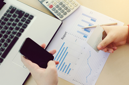 top view of hand holding smart phone and paying with credit card with laptop, expenses chart report and calculator on desk at home office, online payment, shopping online, lifestyle technology conceptの写真素材