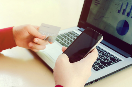 young man holding smart phone and paying with credit card with income and expenses chart report in laptop computer on desk at home office, online payment, shopping online, lifestyle technology conceptの写真素材