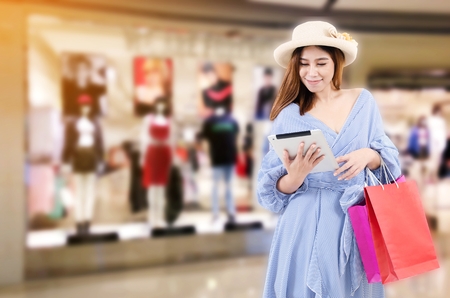 cheerful young asian woman watching special offer in tablet and holding shopping bag while standing at trendy new collection clothes shop in department store shopping mall, shopping online conceptの写真素材