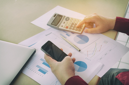 young man hand using smart phone and calculate finance cost with calculator, business strategy diagram report and laptop computer on desk at home office, income and expenses, economy conceptの写真素材