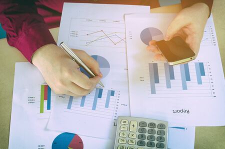 top view of working hand writing and using smart phone with business strategy diagram report and calculator on desk at home office, income and expenses, finance, searching data and economy conceptの写真素材