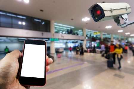 hand using smart phone monitoring and CCTV security indoor camera system operating in airport, people, transportation, surveillance security and safety technology conceptの写真素材
