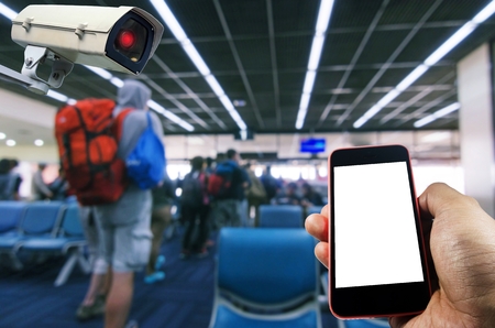 hand using smart phone monitoring and CCTV security indoor camera system operating in airport with passengers waiting at airport, transportation, surveillance security and safety technology conceptの写真素材