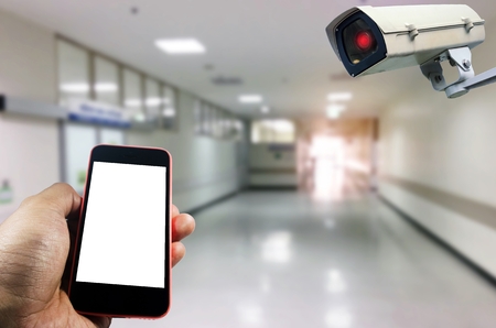 hand using smart phone monitoring and CCTV security indoor camera system operating in hospital background, internet, medical, network connection, surveillance security and safety technology conceptの写真素材