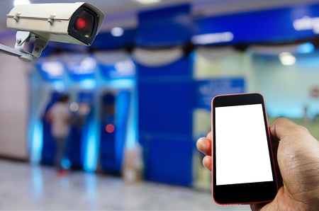 hand holding smart phone monitoring and CCTV security indoor camera system operating with blurred image of people withdraw money from ATM, internet, surveillance security and safety technology conceptの写真素材