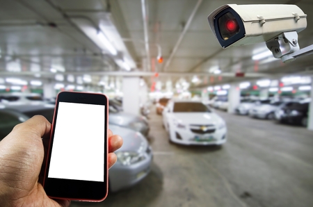 hand holding smart phone monitoring and CCTV security indoor camera system operating with under ground indoor car parking garage area, internet, surveillance security and safety technology conceptの写真素材