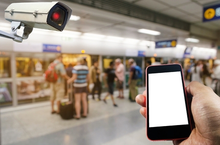 hand holding mobile smart phone and CCTV security indoor camera system operating with people waiting for subway at train station, internet, surveillance security and safety technology conceptの写真素材