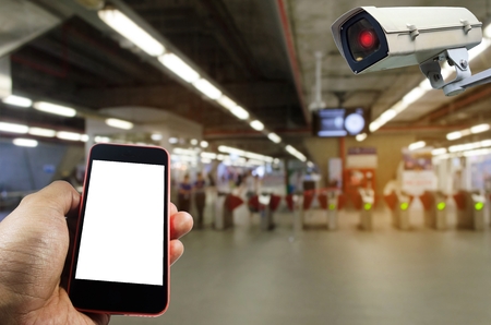 hand holding mobile smart phone and CCTV security indoor camera system operating with blurred image of entrance of subway at train station, internet, surveillance security, safety technology conceptの写真素材