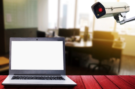 white blank screen laptop computer on wooden desk with CCTV security indoor camera system operating...