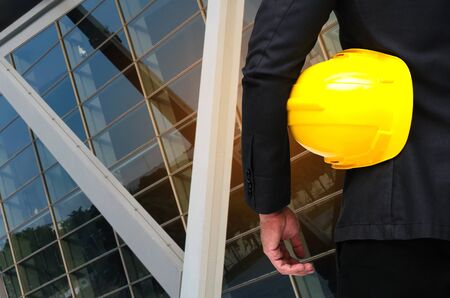 back view of engineer and holding yellow safety helmet standing in front of modern office building background, industrial and business conceptの写真素材