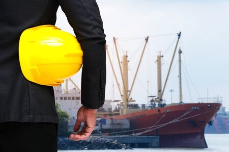 back view of engineer and holding yellow safety helmet standing in front of cargo ship at trade port background, industrial, logistics, transportation and business conceptの写真素材
