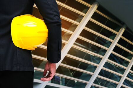 back view of engineer and holding yellow safety helmet standing in front of modern office building...