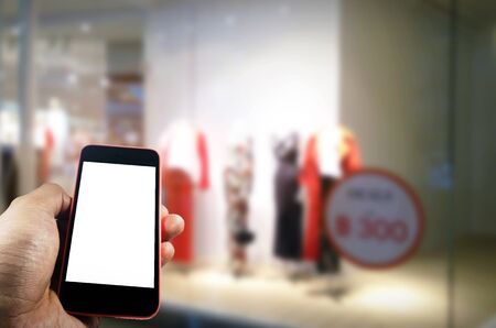 hand using blank screen mobile smart phone with showcases fashion clothes display in department store shopping mall, internet, searching information, social media, payment and shopping online conceptの写真素材