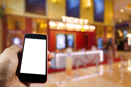 hand using blank screen mobile smart phone to buy cinema tickets with blurred image of ticket sales counter at movie theater, reserved seating, online booking and payment conceptの写真素材