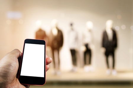 blank screen mobile phone in hand with blurred image of men fashion clothes shop showcase in shopping mall background, internet connection, communication technology and shopping online conceptの写真素材