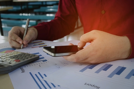 young business man working finance about cost with mobile phone, calculator and strategy diagram chart report on desk at home office, financial, business technology and economy conceptの写真素材