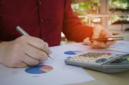 close up young accountant man analysis finance about cost with mobile phone, calculator and business chart on desk at home office, financial, technology, internet, investment and economy conceptの写真素材