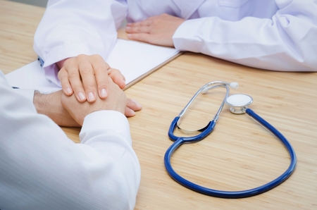 friendly female doctor holding patient hand for reassuring with clipboard and stethoscope on desk in hospital, people, age, health, care and support concept, selective focusの写真素材