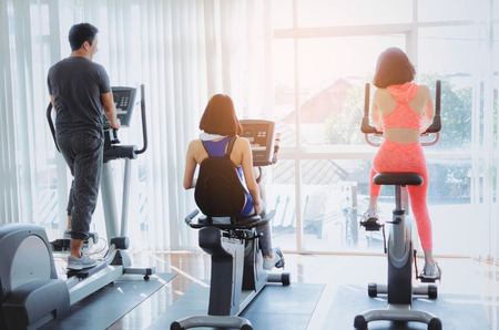 back view of group asian young sportive people doing exercises with treadmill, bicycle machine in...