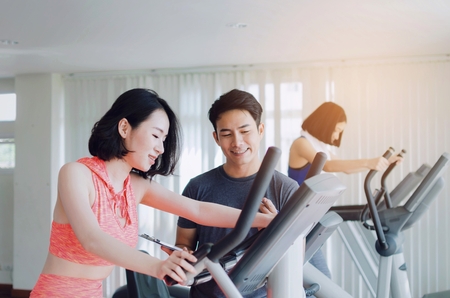asian young woman slim body exercising on bicycle machine with young man personal trainer in fitness gym, bodybuilder, healthy lifestyle, fitness, workout and sport training concept, soft focusの写真素材