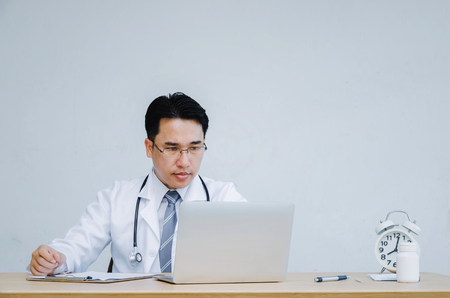 asian doctor with stethoscope around his neck working with laptop computer, alarm clock, pen, pharmacy bottle on desk in hospital, research, science, chemical, health care, medical technology conceptの写真素材