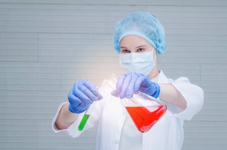 young female scientist in protective mask and gloves dropping red and green liquid substance into...