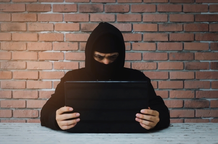 Young Asian man in black jacket with hood stealing information with laptop computer on desk with brick wall background, hacker, crime, network security, programmer and internet technology conceptの写真素材