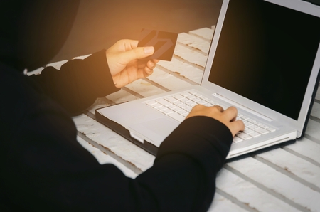 young man in black jacket with hood stealing information with laptop computer and credit card on desk, thief, hacker, crime, network security, programmer, online and internet technology conceptの写真素材