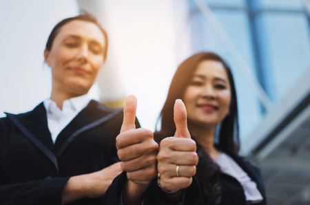 close up hand of two business asian and caucasian woman with mobile phone and giving thumbs up as...
