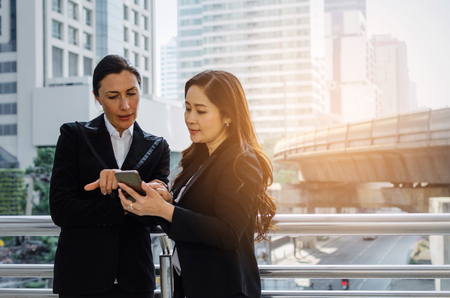 two business asian and caucasian woman talking and reading information about finance news in mobile phone together standing in modern city, network technology, internet, successful, teamwork conceptの写真素材