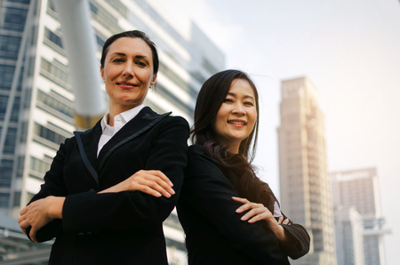 two smiling business asian and caucasian woman in formally dressed looking into camera with her arms crossed standing in modern city, confident, team leader, successful and entrepreneurship conceptの写真素材