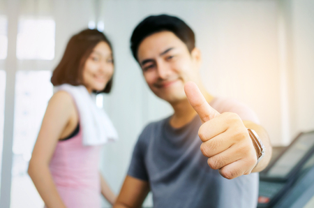 close up hand of muscular handsome man with towel showing thumbs up while resting after workout with blurred young happy smile asian woman slim body background in fitness gym, exercise, sport conceptの写真素材