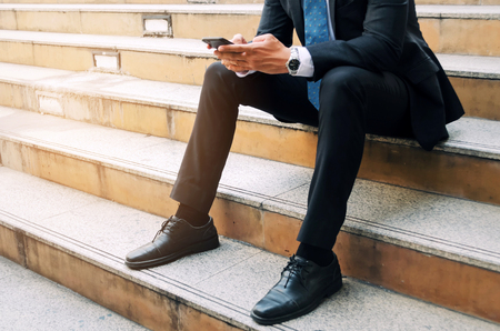 midsection of young business man wearing modern black suit using mobile smart phone and sitting on the walkway in the city, internet network connection, technology communication and financial conceptの写真素材