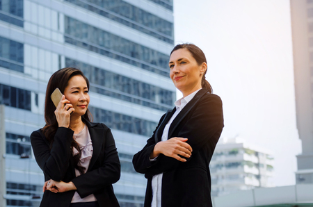 two business asian and caucasian woman with mobile phone and arms crossed standing in the modern city, successful, support, meeting, partner, teamwork, confidence, internet, network connection conceptの写真素材