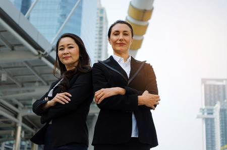 two smiling business asian and caucasian woman in formally dressed looking into camera with her arms crossed standing in modern city, confident, team leader, successful and entrepreneurship conceptの写真素材
